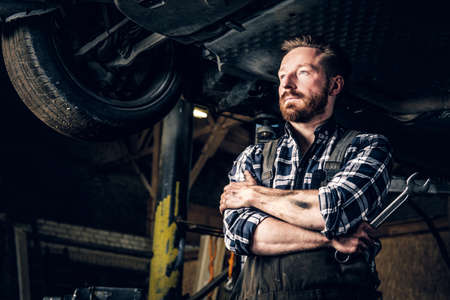 A bearded mechanic with tattoos on arms holds nut key under the car in a garage.の写真素材
