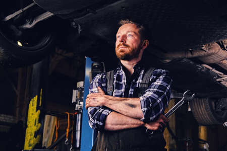 A bearded mechanic with tattoos on arms holds nut key under the car in a garage.の写真素材