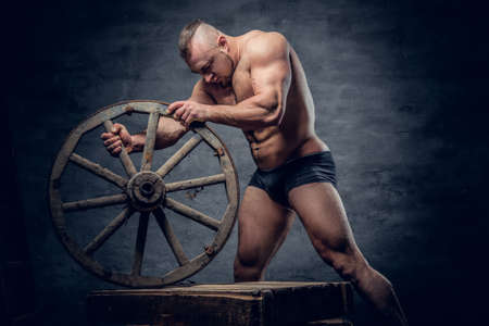 Studio portrait of shirtless muscular bodybuilder slave male dressed in panties holds the old wooden wheel over grey background.の写真素材