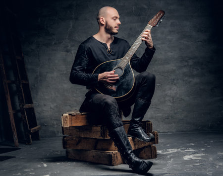Shaved head traditional folk musician dressed in vintage Celtic clothes sits on a wooden box and...