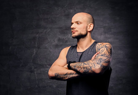 Portrait of shaved head athletic tattooed male with crossed arms over grey background.