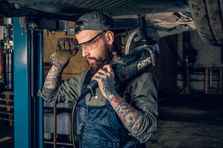 Bearded mechanic in protective googles holds angle grinder on a shoulder under the car in a garage.の写真素材