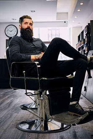 Portrait of a bearded hipster male with stylish haircut sits on a chair in a hairdresser's salon.の写真素材