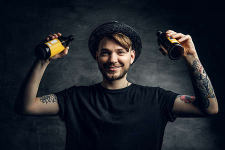 Handsome bearded male with tattooed arms, dressed in a black t shirt and top hat holds craft bottled...