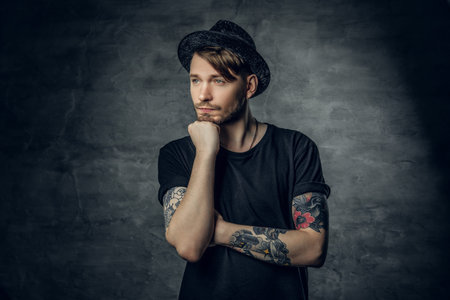 Studio portrait of thoughtful male with crossed tattooed arms, dressed in a black t shirt and tweed...