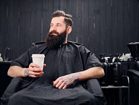 Full throttle bearded hipster male drinks coffee before the hair styling in a hairdresser's salon.の写真素材