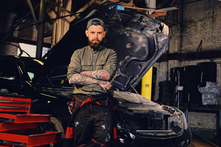 Portrait of bearded tattooed mechanics with crossed arms posing near a car in a workshop.
