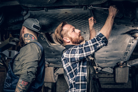 Two bearded tattooed mechanics working under the car in a garage.の写真素材