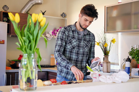 A handsome bearded male dressed in a fleece shirt making salad from vegetables in the home kitchen.の写真素材