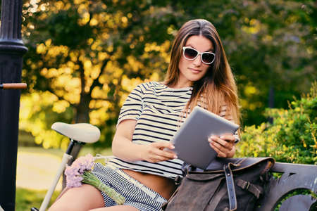 Portrait of brunette female using tablet PC with city bicycle in a park on background.の写真素材