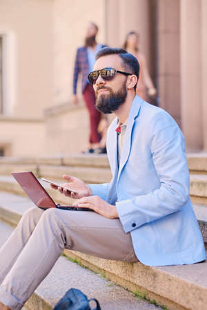 The stylish bearded male sits on a step and using laptop.の写真素材