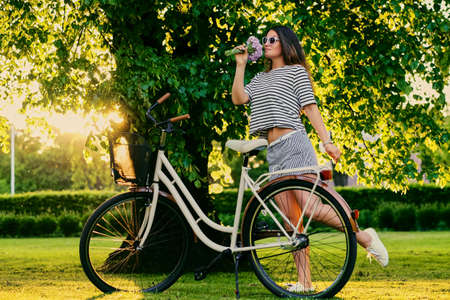 Brunette female holds bouquet near city bicycle in a summer park.の写真素材