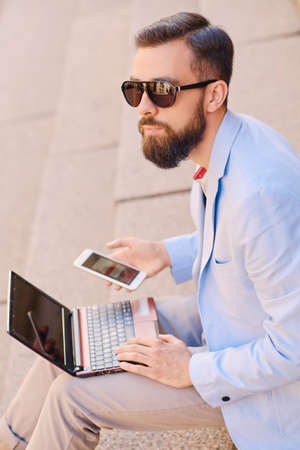 The stylish bearded male sits on a step and using laptop.