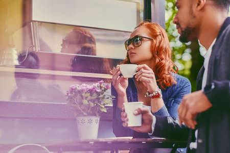 Black bearded male and redhead female drink coffee in a cafe on a street.の写真素材