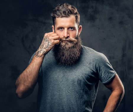 Brutal bearded male with tattoos on arms touches his moustache over grey background.の写真素材