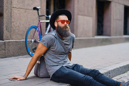 Bearded tattooed male in a hat sits on a step with single speed bicycle on background.の写真素材
