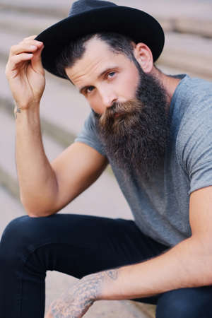 Portrait of a bearded man in a hat with tattoos on arms, sits on a step.の写真素材