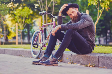 Bearded tattooed male in a hat sits on a step with single speed bicycle on background.の写真素材