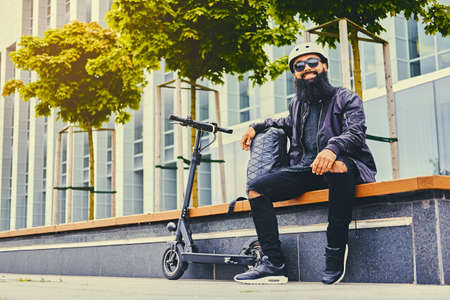 Stylish bearded male in sunglasses relaxing on a bench after riding by electric scooter in downtown.の写真素材