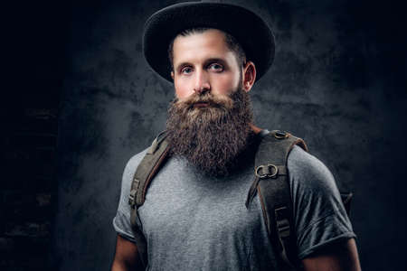 Bearded tattooed male in a hat with a backpack over grey background.
