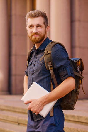 The bearded casual traveler male sits on a step and using a tablet PC.