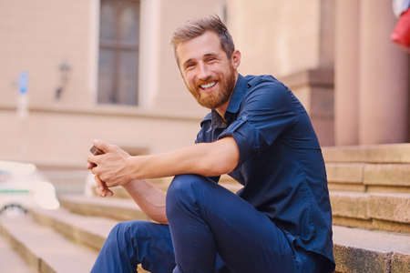 Stylish bearded positive male in a jeans and denim jacket sits on a step and using a smartphone.の写真素材