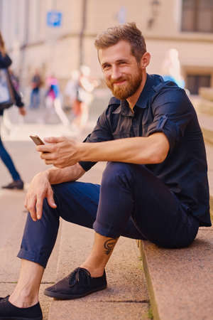 Stylish bearded positive male in a jeans and denim jacket sits on a step and using a smartphone.の写真素材