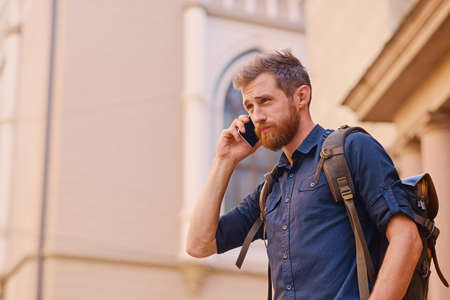 Bearded male with backpack speaking by smart phone in an old European town.の写真素材