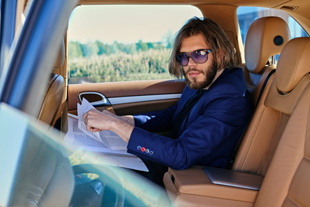 Bearded stylish male sits in a car and working with paper documents.