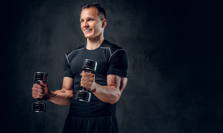 An athletic fitness male dressed in sportswear holds a set of dumbbells over grey background.の写真素材