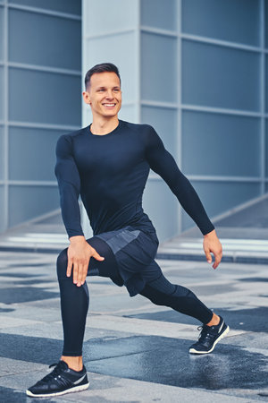 Sporty athletic male is warming up and stretching outdoors over modern building background.