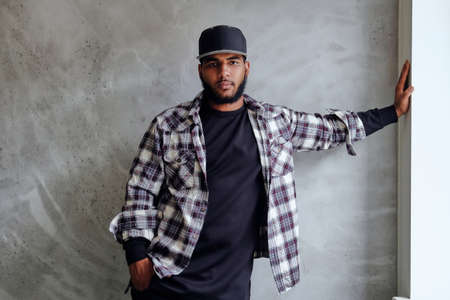 Black, bearded hipster male dressed in a fleece shirt and a cap, posing near window with natural light.の写真素材