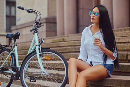 Sensual brunette female in a blue sunglasses sits on a step with city bicycle on background.の写真素材