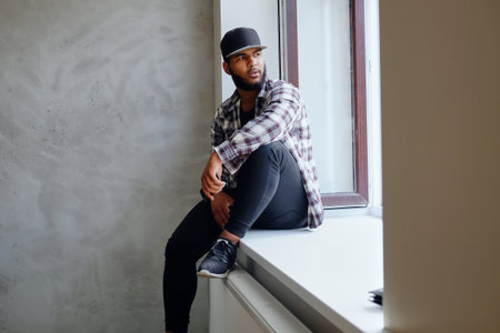 Black, bearded hipster male dressed in a fleece shirt and a cap, posing near window with natural light.の写真素材