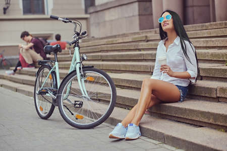 Sensual brunette female in a blue sunglasses sits on a step with city bicycle on background.の写真素材