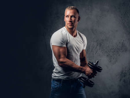 Handsome muscular blond male in a white t shirt holds dumbbell over grey artistic background.の写真素材