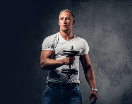 Handsome muscular blond male in a white t shirt holds dumbbell over grey artistic background.の写真素材