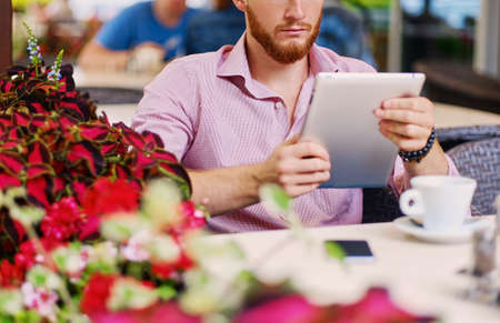 Attractive bearded redhead male in a pink shirt sits at the table in a cafe and using a tablet PC.