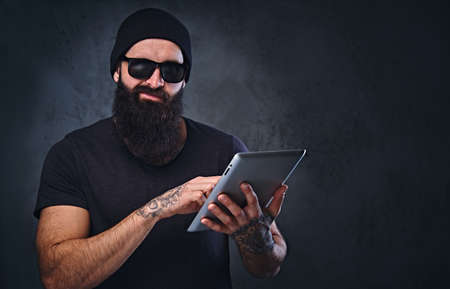 Bearded brutal male in a hat and sunglasses holds tablet PC over grey background.の写真素材