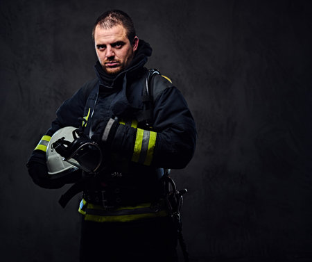 Studio portrait of firefighter dressed in uniform holds safety helmet in his arm.