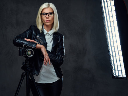 A blond photographer female dressed in a black leather jacket holds professional digital camera and...