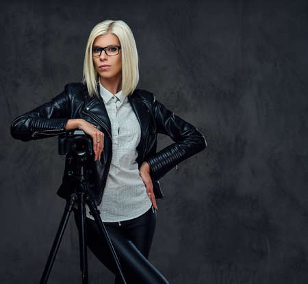 A blond photographer female dressed in a black leather jacket holds professional digital camera and a tripod.の写真素材