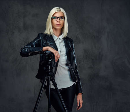 A blond photographer female dressed in a black leather jacket holds professional digital camera and a tripod.の写真素材