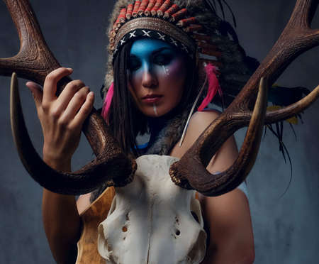 Portrait of shamanic female with Indian feather hat and colorful makeup holds an antler skull over grey background.の写真素材