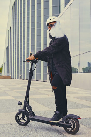 Bearded hipster male vaping and riding by electric scooter over modern building.