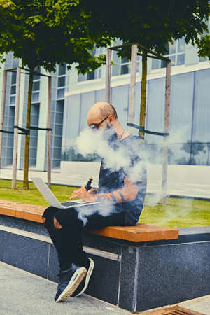 Tattooed bearded, shaved head male using a laptop over modern building background.の写真素材