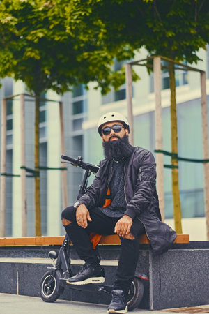 Stylish bearded male in sunglasses relaxing on a bench after riding by electric scooter in downtown.