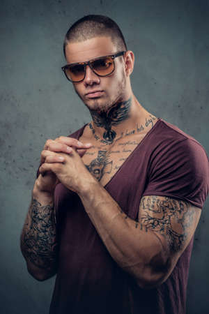 Handsome, athletic male in sunglasses with tattoos on his arms and neck posing over grey artistic background in a studio.の写真素材