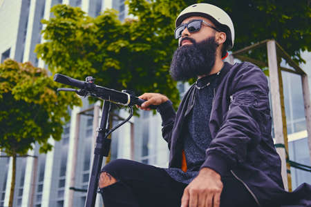 Stylish bearded male in sunglasses relaxing on a bench after riding by electric scooter in downtown.の写真素材