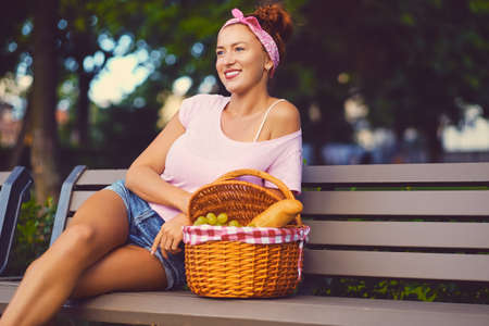 Positive redhead female sits on a bench with a picnic basket full of fruits, bread and wine.の写真素材